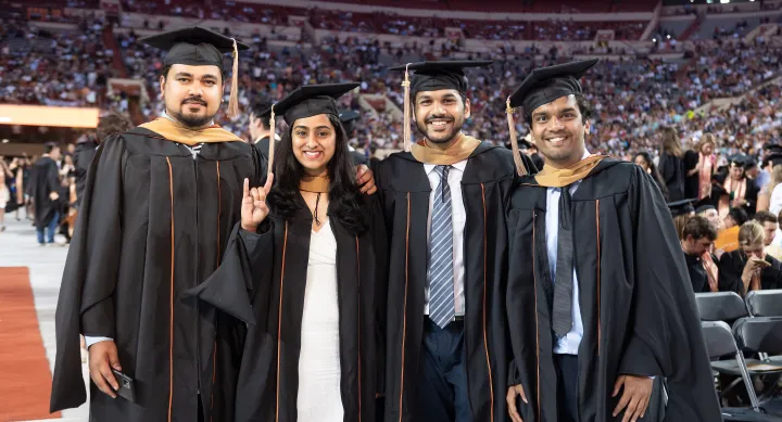 Group of four students, one making the hook 'em gesture, wearing master's regalia